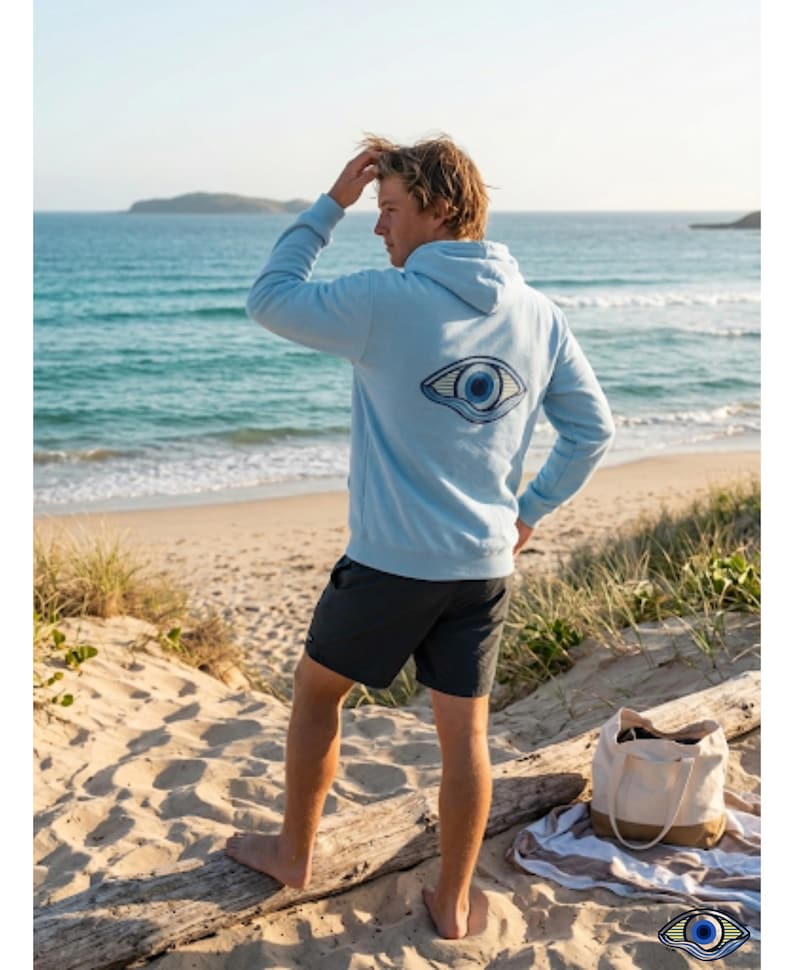 May include: A light blue hoodie with a large eye design on the back. The hoodie is worn with dark gray shorts. A beach bag and towel are on the sand. The person is standing on a sandy beach, looking out at the ocean.