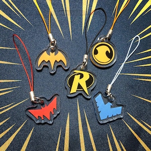 Puede incluir: Cinco dijes de acrílico con logotipos de superhéroes: un murciélago amarillo, un murciélago rojo, una R de Robin amarilla, un círculo de Robin amarillo y un emblema de pájaro azul. Cada dije está unido a un cordón fino de color.