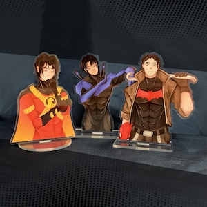 Peut inclure: Trois figurines en acrylique représentant des personnages illustrés. La figurine de gauche porte une tenue rouge et jaune avec une cape. Celle du centre porte une tenue noire et bleue avec deux bâtons. Celle de droite porte une tenue noire avec un emblème rouge sur la poitrine et une veste marron.