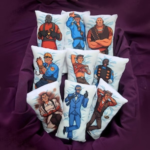 May include: A collection of nine rectangular plush pillows featuring colorful cartoon illustrations of characters from the video game Team Fortress 2, including the Pyro, Soldier, Heavy, Engineer, Scout, Demoman, Medic, Spy, and Sniper.