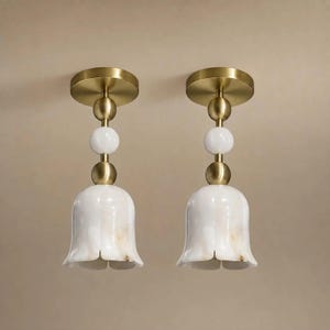 Op de afbeelding: Twee plafondlampen met een ronde messing plafondkap, een verticale stang met twee messing bollen en één witte bol, en een klokvormige witte marmeren kap met subtiele aders. Het ontwerp is symmetrisch en verticaal georiënteerd.
