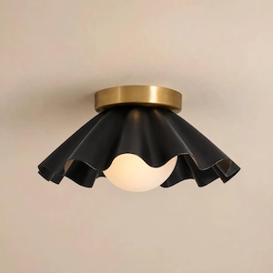 May include: A flush mount ceiling light fixture featuring a matte black, wide, ruffled metal shade. The fixture includes a cylindrical brass-toned base and a central, spherical, frosted white glass globe that emits a soft, diffused light.