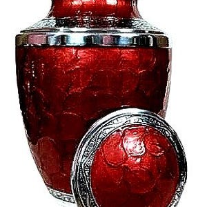 May include: A red cremation urn with a silver-colored rim and lid. The urn has a textured surface and a matching lid, which is placed next to the urn. The urn is designed to hold the ashes of a loved one.