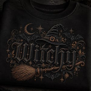 May include: A black crewneck sweatshirt featuring an embroidered design with the word Witchy in gothic lettering. The graphic includes a pointed witch hat, a broomstick, a crescent moon, stars, and bats, all rendered in shades of bronze and gold.