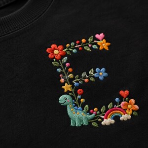 Puede incluir: Una sudadera negra con la letra E bordada formada por flores coloridas, hojas verdes, un pequeño dinosaurio verde claro, una estrella amarilla, un arcoíris diminuto y un corazón rosa. El bordado utiliza hilos vibrantes en rojo, naranja, amarillo, azul y rosa.
