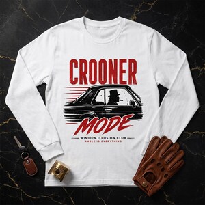 Op de afbeelding: Een wit shirt met lange mouwen en een rode grafische print met de tekst CROONER MODE, WINDOW ILLUSION CLUB en ANGLE IS EVERYTHING. Het ontwerp toont het silhouet van een zwarte auto met een bestuurder die een hoge hoed draagt.