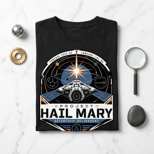 Puede incluir: Una camiseta negra con un estampado gráfico de una nave espacial en el espacio con una estrella brillante, planetas y diagramas científicos. El texto dice "SOMEONE SAVE US SOMEONE LIKE ME", "PROJECT HAIL MARY" y "SCIENTIFIC DELINQUENT" con un símbolo lambda.