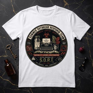 Può includere: Una t-shirt bianca con una grafica circolare che riporta il testo SUMMER OUTBREAK RESPONSE TEAM. Il design include una macchina da scrivere, una fiala etichettata VIRAL SAMPLE, una pianta in vaso e il testo TACTICAL BIOLOGICAL UNIT S.O.R.T. EST. 1996.