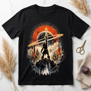 Op de afbeelding: Een zwart t-shirt met een grafische print van een silhouetfiguur die op een rotsachtige bergtop staat en een gloeiend zwaard omhoog houdt tegen een achtergrond van een grote oranje zon, een planeet met ringen en witte wolken.