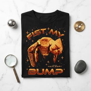 Op de afbeelding: Een zwart t-shirt met een afbeelding van een rotsachtig, meerledig wezen in een fist-bump pose. De tekst FIST MY BUMP en PROJECT HAIL MARY staat in oranje, omringd door kleine sterren op de stof van het t-shirt.