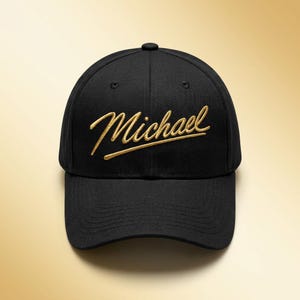 Op de afbeelding: Zwarte baseballcap met de naam "Michael" in gouden script geborduurd op de voorkant. De cap heeft een gebogen klep en een knoop aan de bovenkant. Een stijlvol en gepersonaliseerd accessoire.