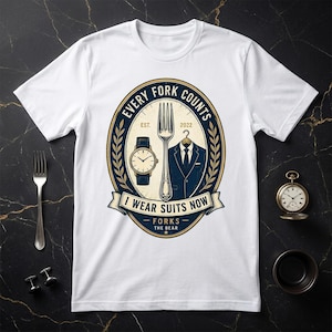 May include: A white t-shirt featuring a central graphic inside an oval frame. The design includes a fork, a wristwatch, and a suit jacket on a hanger, with the text "EVERY FORK COUNTS", "EST. 2022", "I WEAR SUITS NOW", and "FORKS THE BEAR".