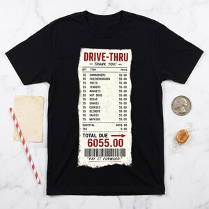 Op de afbeelding: Een zwart t-shirt met korte mouwen en een grafische print van een lange drive-thru kassabon. De bon bevat diverse etenswaren zoals hamburgers en friet met een totaalbedrag van 6055.00 en de tekst "PAY IT FORWARD" boven een streepjescode.