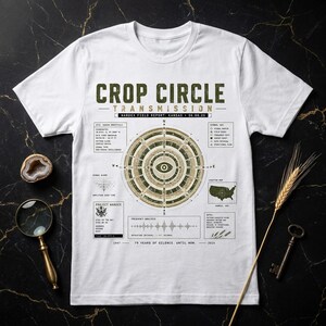 Può includere: Una t-shirt bianca a maniche corte con una stampa grafica intitolata CROP CIRCLE TRANSMISSION. Il design include un motivo circolare centrale, una mappa del Kansas, un'onda di frequenza e un testo tecnico etichettato WARDEX FIELD REPORT: KANSAS 06.06.25.