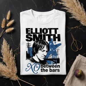 Può includere: Una t-shirt bianca con un ritratto in bianco e nero di Elliott Smith. La grafica include il testo ELLIOTT SMITH, XO e between the bars, insieme a una sagoma blu di un ponte e una stella su uno sfondo urbano nero.