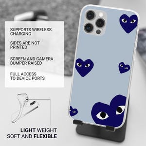 Op de afbeelding: Een doorzichtige telefoonhoes met een lichtblauwe achtergrond en marineblauwe hartvormige oogontwerpen. De hoes staat op een zwarte standaard. Tekst links: Ondersteunt draadloos opladen, zijkanten zijn niet bedrukt, scherm- en camerabumper verhoogd, etc.