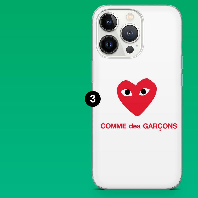 Op de afbeelding: Witte telefoonhoes met een rood hartlogo en de tekst "COMME des GAR&Ccedil;ONS". De hoes heeft een camera-opening en is ontworpen voor een mobiele telefoon.