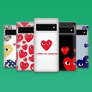 Op de afbeelding: Een verzameling telefoonhoesjes met diverse ontwerpen. E&eacute;n hoesje is wit met een rood hart en de tekst "Comme des Gar&ccedil;ons". Andere hoesjes hebben herhaalde hartpatronen, camouflage en een zwart hoesje met een rood hart. De hoesjes staan tegen een groene achtergrond.
