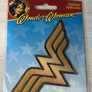 May include: A gold embroidered Wonder Woman logo appliqué patch on a blue and red backing card. The patch features the iconic double-W emblem with black stitched borders. The packaging includes the text Wonder Woman and Appliqué Aplicación.