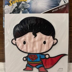 May include: A Superman embroidered iron-on patch featuring a stylized cartoon character with black hair, a blue suit, a red cape, and a yellow belt. The patch is packaged in a clear plastic sleeve with a header card displaying the Superman logo.