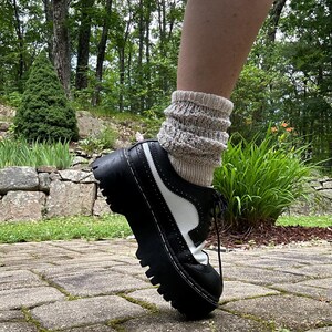 May include: A black and white leather oxford shoe with a thick, lugged black platform sole and brogue detailing. The shoe is paired with a beige, textured, speckled knit sock that reaches above the ankle, featuring a ribbed cuff design.