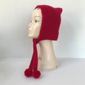 May include: Red knitted hat with a long pointed hood and two pom-poms on the ends of the ties.