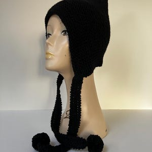May include: Black knitted hat with ear flaps and pom poms. The hat has a pointed top and is made of a soft, warm yarn.