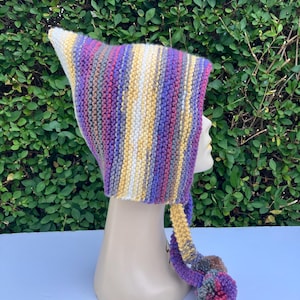 May include: A hand-knitted pointed hat featuring vertical stripes in shades of purple, yellow, white, and grey. The hat has a distinct triangular peak and includes two long braided ties hanging from the sides, each finished with a multi-coloured yarn pom-pom.