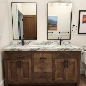 May include: A double bathroom vanity with a white marble countertop featuring gray veining, two rectangular sinks, and dark wood cabinetry. The unit includes two lower cabinets with doors and three central drawers, all fitted with matte black hardware.