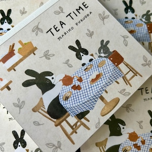 Peut inclure: Une couverture de livre carrée intitulée TEA TIME MARIKO FUKUOKA illustrée de lapins noirs et blancs assis à une table avec une nappe à carreaux bleus et blancs, une théière brune et des petits gâteaux, entourés de petits motifs de feuilles.