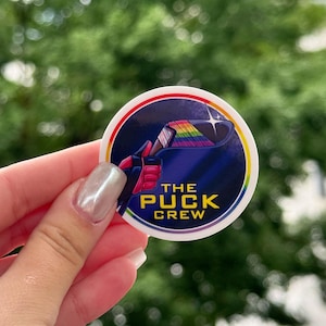 May include: A round sticker with a hockey-themed design. The sticker features a hockey stick and a rainbow-colored puck, with the text "THE PUCK CREW" in white. The sticker has a white border with a rainbow-colored edge.