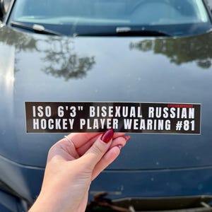 Könnte beinhalten: Ein schwarzer rechteckiger Aufkleber mit weißem Text: "ISO 6'3" BISEXUELLER RUSSISCHER HOCKEYSPIELER MIT DER RÜCKENNUMMER #81." Der Aufkleber wird vor einem schwarzen Auto gehalten.