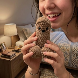 May include: A brown, phallic-shaped crochet plush toy made of soft, textured yarn. The plush features two round base sections, a cylindrical body, and a rounded top. It has small black plastic safety eyes, a stitched black mouth, and pink blush.