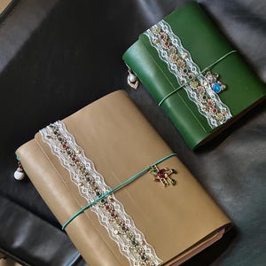 Può includere: Due diari in pelle, uno color cuoio e uno verde scuro, ciascuno caratterizzato da una striscia di pizzo bianco decorata con strass multicolori. Un elastico turchese chiude ogni diario, arricchito da un ciondolo pendente e una perlina bianca.