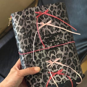 Könnte beinhalten: Ein rechteckiger Notizbuchumschlag mit einem grauen und schwarzen Leopardenmuster. Der Umschlag ist mit mehreren Ösen verziert und mit dünnen Bändern in den Farben Pink, Hellrosa und Schwarz geschnürt, die vorne zu kleinen Schleifen gebunden sind.