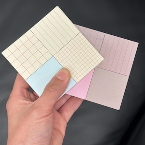 Könnte beinhalten: Zwei quadratische Notizblöcke mit Raster- und Linienmustern. Ein Notizblock hat ein hellblaues Quadrat, der andere ein rosa Quadrat. Die Notizblöcke werden in einer Hand vor einem schwarzen Hintergrund gehalten.