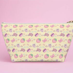 Könnte beinhalten: Eine trapezförmige Reißverschlusstasche mit hellgelbem Hintergrund und einem wiederkehrenden Muster aus rosa Erdbeeren, lila Macarons, rosa Schleifen, kleinen blauen Herzen und rosa Kameras mit einem goldfarbenen Reißverschlussanhänger oben rechts.