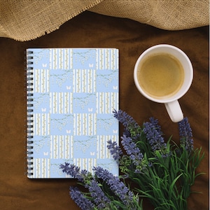 Puede incluir: Un cuaderno de espiral con una cubierta azul claro que presenta un patrón repetitivo de rayas verticales blancas, delicadas ramas florales colgantes y pequeñas mariposas blancas, junto a una taza de café y lavanda morada.