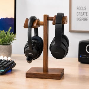 Könnte beinhalten: Ein Kopfhörerständer aus dunkelbraunem Holz mit rechteckigem Sockel und einem zentralen vertikalen Pfosten mit zwei horizontalen Armen. Zwei schwarze Bose-Over-Ear-Kopfhörer hängen an den Armen des Ständers, einer auf jeder Seite des Pfostens.
