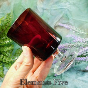 May include: A dark red glass wine goblet with a clear glass stem and base. The goblet is being held by a hand.