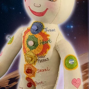 K&ouml;nnte beinhalten: Handgefertigte Puppe mit l&auml;chelndem Gesicht und bemalten Chakra-Symbolen auf dem Torso. Die Puppe hat ein drittes Auge und verschiedene Edelsteinakzente. Der Hintergrund zeigt eine kosmische Szene mit einem Planeten und Sternen.