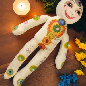 K&ouml;nnte beinhalten: Eine skurrile, cremefarbene Puppe mit bemalten Gesichtsz&uuml;gen und farbenfrohen Chakra-Symbolen. Die Puppe hat ein l&auml;chelndes Gesicht, und ihr K&ouml;rper ist mit verschiedenen Edelsteinen und dekorativen Elementen verziert. Die Puppe ist von einer brennenden Kerze, Blumen und Gr&uuml;n umgeben.