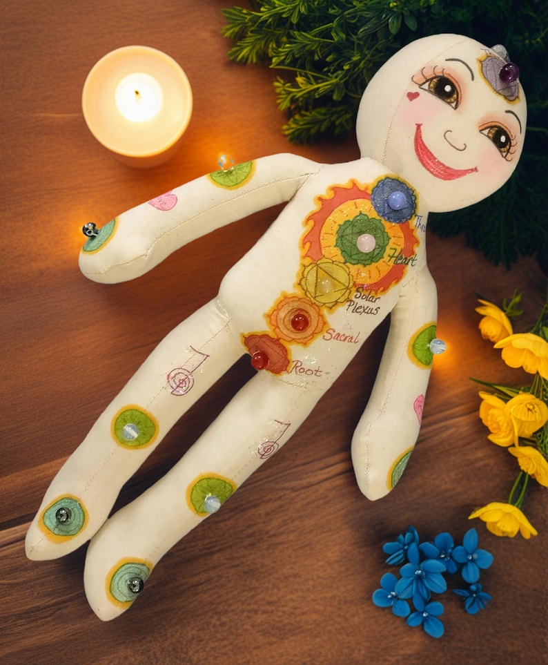 K&ouml;nnte beinhalten: Eine skurrile, cremefarbene Puppe mit bemalten Gesichtsz&uuml;gen und farbenfrohen Chakra-Symbolen. Die Puppe hat ein l&auml;chelndes Gesicht, und ihr K&ouml;rper ist mit verschiedenen Edelsteinen und dekorativen Elementen verziert. Die Puppe ist von einer brennenden Kerze, Blumen und Gr&uuml;n umgeben.