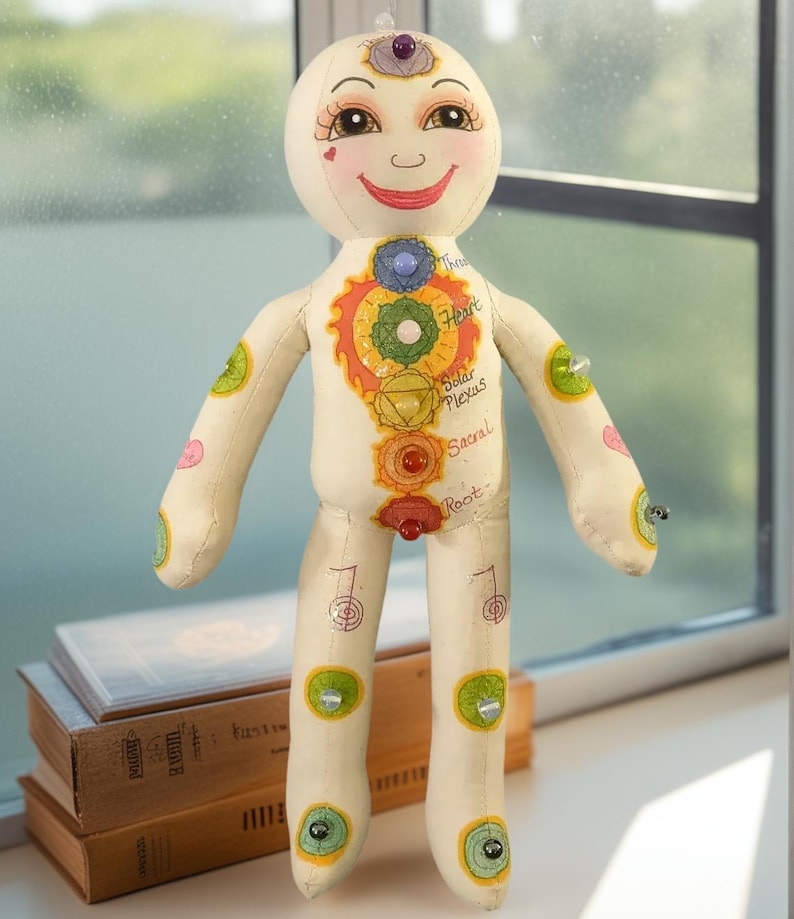 K&ouml;nnte beinhalten: Eine cremefarbene Stoffpuppe mit einem l&auml;chelnden Gesicht und bemalten Merkmalen. Die Puppe hat eine Reihe farbenfroher Chakra-Symbole entlang der K&ouml;rpermitte. Die Puppe hat einen lila Edelstein auf dem Kopf und ist mit verschiedenfarbigen Edelsteinen verziert.