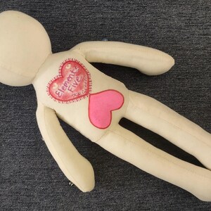 K&ouml;nnte beinhalten: Eine cremefarbene Stoffpuppe mit rundem Kopf und langen Gliedma&szlig;en. Die Puppe hat ein herzf&ouml;rmiges Design auf der Brust, mit den Worten "Elements Five" in einem roten Herzen und einem rosa Herzen darunter.