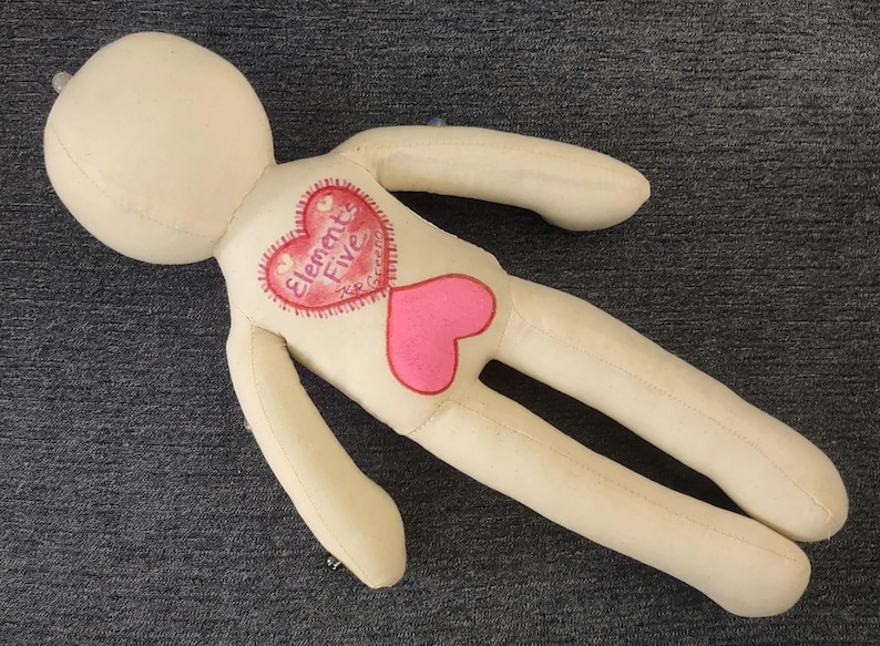 K&ouml;nnte beinhalten: Eine cremefarbene Stoffpuppe mit rundem Kopf und langen Gliedma&szlig;en. Die Puppe hat ein herzf&ouml;rmiges Design auf der Brust, mit den Worten "Elements Five" in einem roten Herzen und einem rosa Herzen darunter.