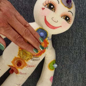 K&ouml;nnte beinhalten: Eine skurrile, handgefertigte Puppe mit einem l&auml;chelnden Gesicht, bemalten Merkmalen und farbenfrohen Chakra-Symbolen. Die Puppe hat einen wei&szlig;en K&ouml;rper mit bemalten Details, einschlie&szlig;lich der W&ouml;rter "Solar Plexus" und "Sacral". Sie hat einen lila Edelstein auf dem Kopf.