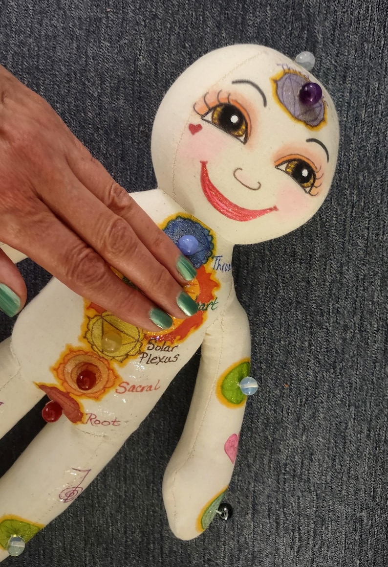 K&ouml;nnte beinhalten: Eine skurrile, handgefertigte Puppe mit einem l&auml;chelnden Gesicht, bemalten Merkmalen und farbenfrohen Chakra-Symbolen. Die Puppe hat einen wei&szlig;en K&ouml;rper mit bemalten Details, einschlie&szlig;lich der W&ouml;rter "Solar Plexus" und "Sacral". Sie hat einen lila Edelstein auf dem Kopf.