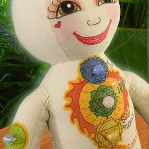 K&ouml;nnte beinhalten: Eine handgefertigte Puppe mit einem l&auml;chelnden Gesicht, bemalten Merkmalen und farbenfrohen Chakra-Symbolen. Die Puppe hat einen cremefarbenen K&ouml;rper mit Edelsteinakzenten und den Worten "Heart", "Solar Plexus", "Sacral" und "Root" darauf geschrieben.