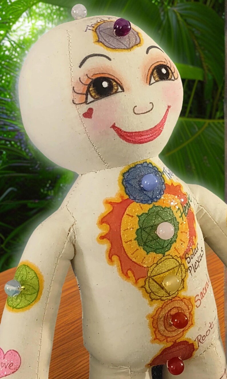 K&ouml;nnte beinhalten: Eine handgefertigte Puppe mit einem l&auml;chelnden Gesicht, bemalten Merkmalen und farbenfrohen Chakra-Symbolen. Die Puppe hat einen cremefarbenen K&ouml;rper mit Edelsteinakzenten und den Worten "Heart", "Solar Plexus", "Sacral" und "Root" darauf geschrieben.